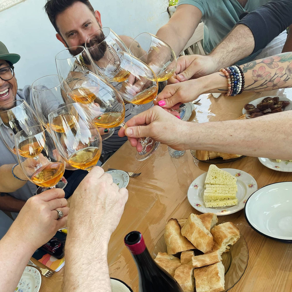 A group of Georgian and American friends raising glasses of amber (orange) wine in a toast. The scene features multiple hands holding wine glasses filled with amber wine, a wooden table with sliced Shotis Puri bread and Imeruli cheese on the table